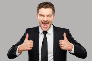 Used to success. Cheerful young man in formalwear showing his thumbs up and smiling while standing against grey background photo