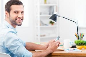 enamorado de mi trabajo. vista lateral de un apuesto joven con camisa sosteniendo un teléfono móvil y sonriendo a la cámara mientras se sienta en su lugar de trabajo foto