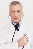 Doctor examining himself. Cheerful grey hair doctor in white uniform examining himself with stethoscope and looking at camera while standing isolated on white photo