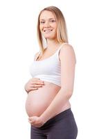 I can feel my baby moving. Side view of beautiful pregnant woman holding hands on her abdomen and smiling while standing isolated on white photo