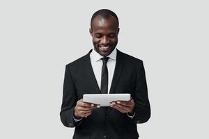 Charming young African man in formalwear working using digital tablet while standing against grey background photo