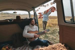 Carefree journey.  Handsome young man playing guitar for his beautiful girlfriend while sitting in retro style mini van photo