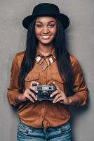 fotógrafo alegre. bella joven africana alegre sosteniendo una cámara de estilo retro y mirando la cámara con una sonrisa mientras se enfrenta a un fondo gris foto