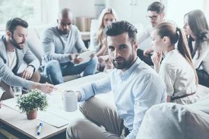Feeling confident in my team. Group of confident business people sitting at the office desk together and discussing something while young man holding coffee cup and looking over shoulder photo