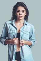 demasiado sexy para su camisa. atractiva mujer joven en lencería quitándose la camisa de jeans y mirando a la cámara mientras está de pie contra el fondo gris foto