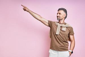 Handsome mature man pointing away and smiling while standing against pink background photo