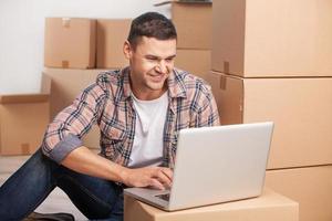 Surfing web in new apartment. Cheerful young man sitting on the floor and working on laptop while cardboard boxes laying on the background photo