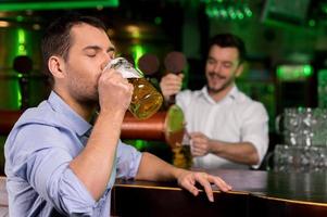 Drinking a freshly tapped beer. Handsome young man drinking beer while bartender tapping beer on the background photo