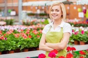 I love to work with flowers. Beautiful young woman in apron keeping arms crossed and smiling while standing in a greenhouse with flowers all around her photo