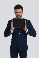 Copy space on his tablet. Good looking young man in full suit holding digital tablet and looking at camera while standing against grey background photo