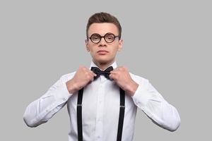 Confident in his skills. Serious young nerd man adjusting his bow tie and looking at camera while standing against grey background photo