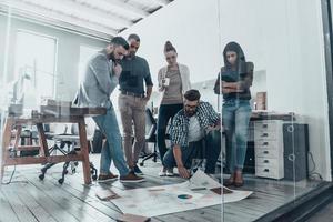 Discussing strategy. Handsome young man with beard pointing at large paper laying on floor while his colleagues looking at it photo