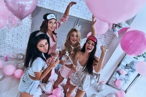 vista superior de la fiesta de damas de cuatro atractivas jóvenes sonrientes en pijama bebiendo champán mientras tienen una fiesta de pijamas en el dormitorio foto