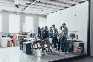 el trabajo en equipo es la clave del éxito. toda la longitud de los jóvenes modernos con ropa informal elegante discutiendo negocios mientras están de pie detrás de la pared de vidrio en la oficina creativa foto