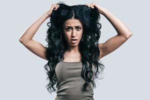 Confused with a new hairstyle. Attractive young woman in casual wear holding hands in hair with confused face while standing against grey background photo