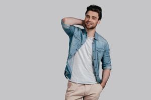 Cheerful and handsome. Good looking young man in blue jeans shirt holding hand behind head and looking at camera while standing against grey background photo