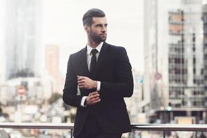 Modern businessman. Confident young man in full suit adjusting his sleeve and looking away while standing outdoors with cityscape in the background photo