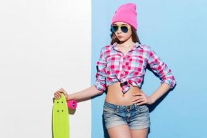 Confident skater girl. Beautiful young woman in pink headwear and sunglasses leaning skateboard while standing against colorful background photo