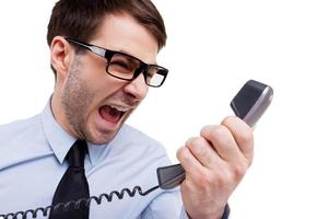 Stressful office life. Furious young man in shirt and tie shouting at telephone while standing isolated on white photo