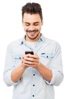 Nowhere without my phone. Handsome young man holding mobile phone and smiling while standing against white background photo