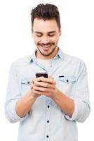 Nowhere without my phone. Handsome young man holding mobile phone and smiling while standing against white background photo