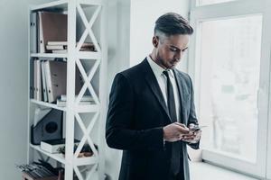 Quick message. Good looking young man in full suit using his smart phone while standing near the window in the office photo