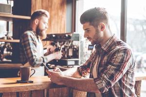 Spending time at coffee shop. Side view of young handsome man using his smartphone while sitting at bar counter at cafe with barista at the background photo