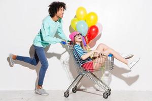 pasar un buen rato juntos. un joven feliz cargando a su bella novia en un carrito de compras y sonriendo mientras corre contra un fondo gris foto