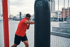 un joven fuerte con ropa deportiva pateando un saco de boxeo mientras hace ejercicio al aire libre foto