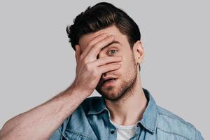 What is there Handsome young man in blue jeans shirt covering face with hand and looking at camera while standing against grey background photo