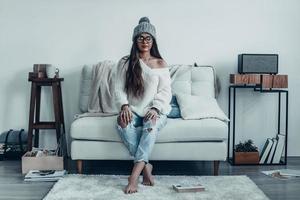 Fashionable girl.  Beautiful young woman in casual wear and knit hat looking at camera while sitting on the sofa at home photo