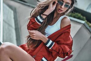 Young and cool. Attractive young woman in sport clothing adjusting her sunglasses while sitting outdoors photo