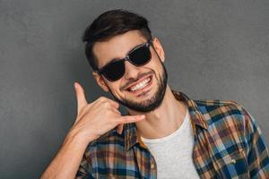 Call me Portrait of cheerful young man gesturing telephone with his hand and looking at camera with smile while standing against grey background photo