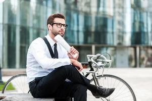 Quick break during busy day. Side view of thoughtful young businessman holding newspaper and looking away while sitting near his bicycle with office building in the background photo