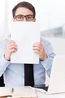 cansado del papeleo. sorprendido joven con camisa y corbata sosteniendo papel y mirando hacia afuera mientras estaba sentado en su lugar de trabajo foto