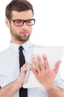 Using opportunities of digital age. Cheerful young man in formalwear and glasses working on digital tablet while standing isolated on white background photo