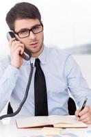 It is so much work to do. Confident young man in shirt and tie talking on the mobile phone and looking at camera while sitting at his working place photo