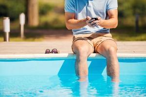 Chatting with friends. Cropped image of man sitting poolside and typing something on mobile phone photo