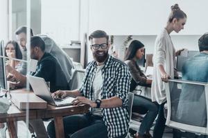 Happy to be a part of great team.  Young handsome man with beard smiling and looking at camera while sitting in office with colleagues working in the background photo