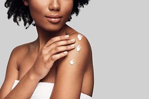 Beauty routine. Close up of young African woman applying body cream and smiling while standing against grey background photo