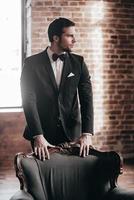 Confidence and contemplation. Thoughtful young handsome man in full suit and bow tie leaning at the chair and looking away while standing in loft interior photo