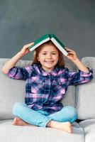 Lets play hide and seek Cheerful little girl holding book over her head and looking at camera with smile while sitting on the couch in lotus position at home photo