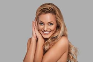 Nice young woman with fresh glowing skin looking at camera and smiling while standing against grey background photo