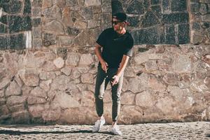 Young and carefree. Full length of handsome young African man in casual clothes moving and gesturing while standing against the stoned wall outdoors photo