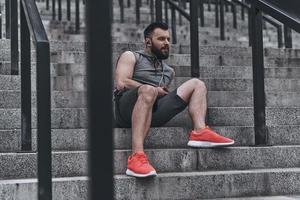 Enjoying music after good run. Handsome young man in sport clothing listening music while sitting on the steps outside photo