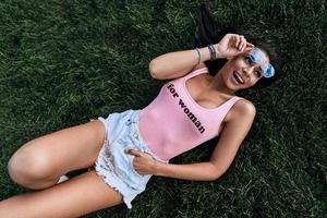 Moments of happiness. Top view of attractive young woman in swimwear and shorts adjusting her eyewear and smiling while lying down on the grass photo