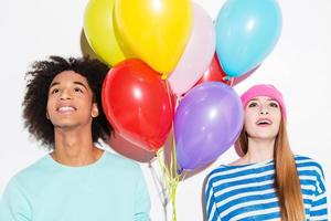 In their own colorful world. Funky young couple holding balloons and looking up while standing against white background photo