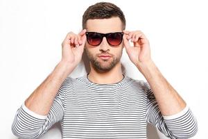 Handsome and in style. Handsome young man adjusting his sunglasses and looking at camera while standing against white background photo