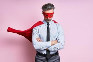 Confident mature man in shirt and tie wearing superhero cape while standing against pink background photo
