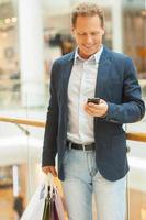 Man shopping. Cheerful mature man holding shopping bags and looking at his mobile phone while standing in shopping mall photo